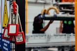 Several red padlocks with 'Locked Out Do Not Remove' tags hang from a metal bar in a manufacturing or industrial setting. Blurry figures are seen working on machinery in the background, emphasizing a safety protocol in place.