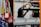 Several red padlocks with 'Locked Out Do Not Remove' tags hang from a metal bar in a manufacturing or industrial setting. Blurry figures are seen working on machinery in the background, emphasizing a safety protocol in place.
