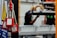 Several red padlocks with 'Locked Out Do Not Remove' tags hang from a metal bar in a manufacturing or industrial setting. Blurry figures are seen working on machinery in the background, emphasizing a safety protocol in place.