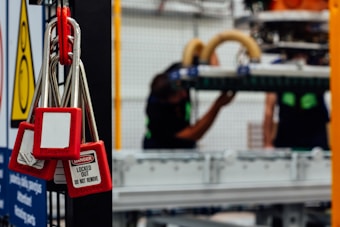Several red padlocks with 'Locked Out Do Not Remove' tags hang from a metal bar in a manufacturing or industrial setting. Blurry figures are seen working on machinery in the background, emphasizing a safety protocol in place.