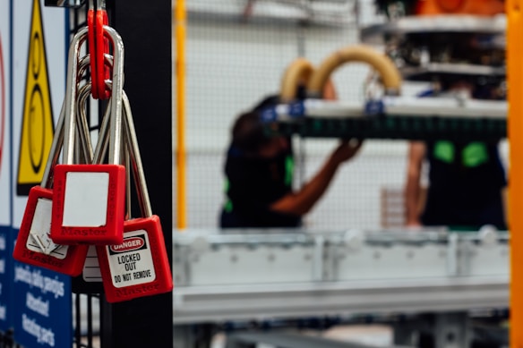 Several red padlocks with 'Locked Out Do Not Remove' tags hang from a metal bar in a manufacturing or industrial setting. Blurry figures are seen working on machinery in the background, emphasizing a safety protocol in place.