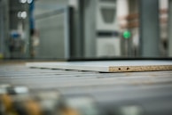 A close-up view of a piece of particle board with two visible holes on its side, placed on a metal conveyor belt within an industrial setting. The background is blurred, highlighting machinery and equipment.