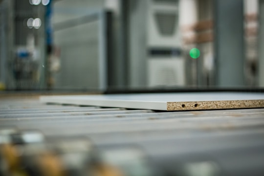 A close-up view of a piece of particle board with two visible holes on its side, placed on a metal conveyor belt within an industrial setting. The background is blurred, highlighting machinery and equipment.