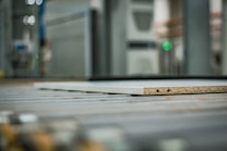 A close-up view of a piece of particle board with two visible holes on its side, placed on a metal conveyor belt within an industrial setting. The background is blurred, highlighting machinery and equipment.