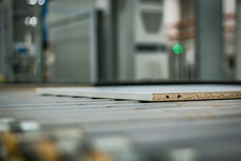 A close-up view of a piece of particle board with two visible holes on its side, placed on a metal conveyor belt within an industrial setting. The background is blurred, highlighting machinery and equipment.