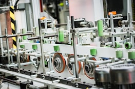 A complex arrangement of machinery features metal rods and various colored components. Green cylinders are attached to the structure alongside orange wheels and metal frames. The machinery appears to be part of an industrial or manufacturing process.