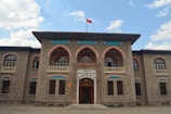 A historic stone building with arched windows and ornate architectural details. A Turkish flag flies atop the structure, and there is an entrance with a sign above it. The sky in the background is partly cloudy.
