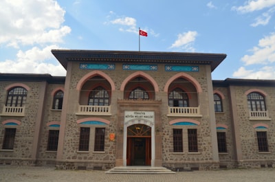 A historic stone building with arched windows and ornate architectural details. A Turkish flag flies atop the structure, and there is an entrance with a sign above it. The sky in the background is partly cloudy.