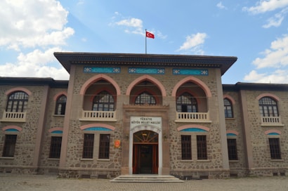 A historic stone building with arched windows and ornate architectural details. A Turkish flag flies atop the structure, and there is an entrance with a sign above it. The sky in the background is partly cloudy.
