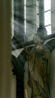 A detailed statue of a religious figure, possibly a saint, holding a globe with a cross. Sunlight streams through a large, stained glass window, illuminating the stone facade of the church. The architecture includes tall, dark stone pillars and intricate designs, adding to the sacred and historical atmosphere.