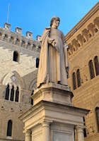 A large stone statue of a figure in historical attire holding a book stands on a pedestal. The background includes an ornate architectural facade with Gothic and Romanesque elements, featuring arched windows and decorative stonework. The clear blue sky provides a striking contrast to the earthy tones of the buildings.