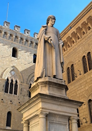 A large stone statue of a figure in historical attire holding a book stands on a pedestal. The background includes an ornate architectural facade with Gothic and Romanesque elements, featuring arched windows and decorative stonework. The clear blue sky provides a striking contrast to the earthy tones of the buildings.
