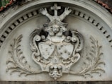 A stone relief sculpture features a central emblem with detailed carvings. At the top, a cross is present with two outstretched arms. Below, a figure's face is visible, and two shields with intricate designs are positioned beneath it. The relief is framed with ornate leaf patterns on both sides and a decorative arch above.
