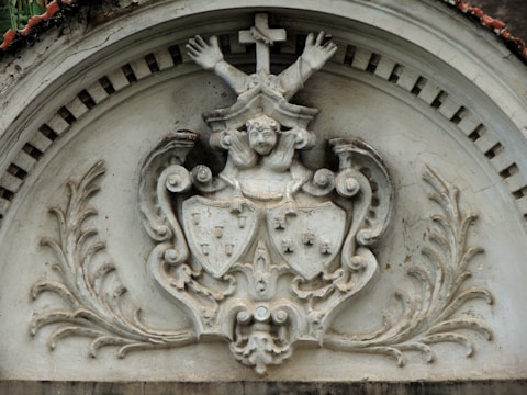 A stone relief sculpture features a central emblem with detailed carvings. At the top, a cross is present with two outstretched arms. Below, a figure's face is visible, and two shields with intricate designs are positioned beneath it. The relief is framed with ornate leaf patterns on both sides and a decorative arch above.