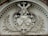 A stone relief sculpture features a central emblem with detailed carvings. At the top, a cross is present with two outstretched arms. Below, a figure's face is visible, and two shields with intricate designs are positioned beneath it. The relief is framed with ornate leaf patterns on both sides and a decorative arch above.