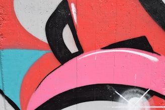 A vibrant graffiti mural features bold, overlapping shapes in pink, red, black, white, and blue. The design includes curved and angular lines with a textured wall surface, giving it a dynamic and energetic feel.
