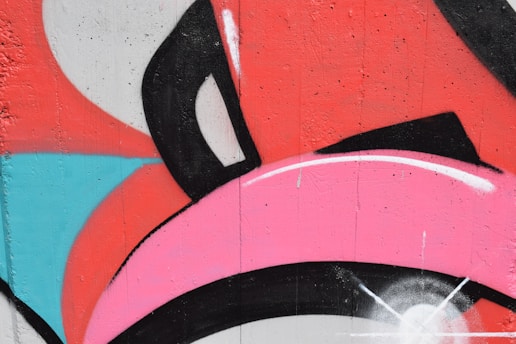 A vibrant graffiti mural features bold, overlapping shapes in pink, red, black, white, and blue. The design includes curved and angular lines with a textured wall surface, giving it a dynamic and energetic feel.