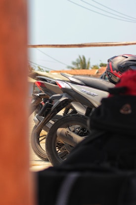A row of parked motorcycles is partially visible, with a close-up view of a motorcycle's rear wheel and tail section. The background features blurred greenery and rooftops, while a helmet rests on one of the bikes, and the scene is framed by wooden posts.