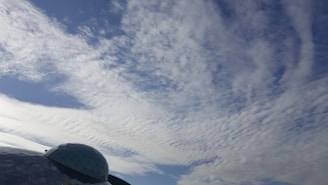 A large portion of the image is dominated by a clear sky with scattered clouds creating a textured pattern. In the lower left corner, there is a geodesic dome structure made of triangular glass panels, reflecting light and partially covered by shadows.