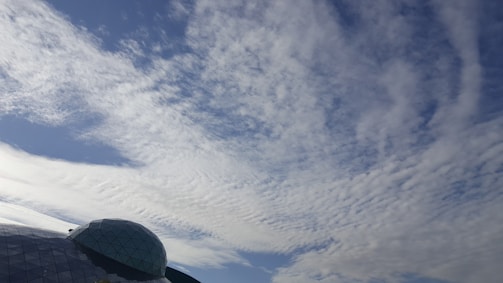 A large portion of the image is dominated by a clear sky with scattered clouds creating a textured pattern. In the lower left corner, there is a geodesic dome structure made of triangular glass panels, reflecting light and partially covered by shadows.