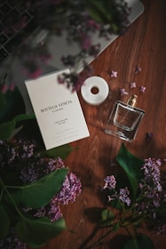 A Bottega Veneta perfume box and bottle are placed on a wooden surface, surrounded by lilac flowers and green leaves. The subtle lighting creates a luxurious and serene atmosphere.