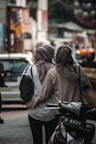 Two people wearing hijabs walk closely together down a bustling urban street. One is carrying a black bag while the other has their arm around the first person. They are surrounded by blurred background elements that suggest a busy market or street scene, with vehicles and colorful objects.