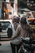 Two people wearing hijabs walk closely together down a bustling urban street. One is carrying a black bag while the other has their arm around the first person. They are surrounded by blurred background elements that suggest a busy market or street scene, with vehicles and colorful objects.