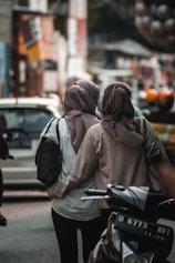 Two people wearing hijabs walk closely together down a bustling urban street. One is carrying a black bag while the other has their arm around the first person. They are surrounded by blurred background elements that suggest a busy market or street scene, with vehicles and colorful objects.