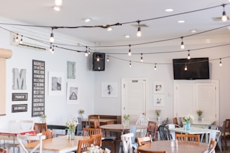 A cozy café interior with wooden tables and a variety of chairs. The walls are adorned with framed monochromatic art and signs listing city names. String lights hang from the ceiling, adding a warm ambiance. Small vases with flowers decorate each table, and a television is mounted on the wall.