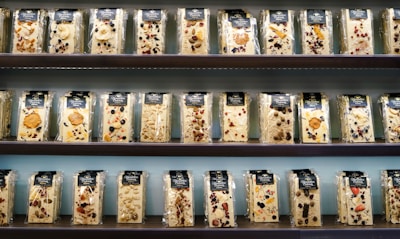 A display of various packaged nougat bars arranged on shelves. Each package contains a nougat bar embedded with assorted nuts, dried fruits, and berries. The bars are wrapped in clear plastic with a dark-colored label at the top, showcasing different combinations of ingredients like almonds, cranberries, and blueberries. The shelves are part of a retail setting with an orderly presentation.