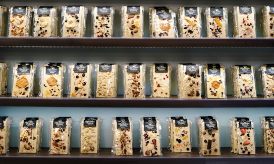 A display of various packaged nougat bars arranged on shelves. Each package contains a nougat bar embedded with assorted nuts, dried fruits, and berries. The bars are wrapped in clear plastic with a dark-colored label at the top, showcasing different combinations of ingredients like almonds, cranberries, and blueberries. The shelves are part of a retail setting with an orderly presentation.