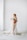 A pregnant woman is standing barefoot in a minimalist room with a white background. She is draped in a flowing white cloth and holding a bouquet of delicate flowers. The cloth partially covers her body while some of it spreads on the floor. She has blonde hair that is styled in waves and is looking off into the distance.