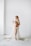 A pregnant woman is standing barefoot in a minimalist room with a white background. She is draped in a flowing white cloth and holding a bouquet of delicate flowers. The cloth partially covers her body while some of it spreads on the floor. She has blonde hair that is styled in waves and is looking off into the distance.