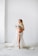 A pregnant woman is standing barefoot in a minimalist room with a white background. She is draped in a flowing white cloth and holding a bouquet of delicate flowers. The cloth partially covers her body while some of it spreads on the floor. She has blonde hair that is styled in waves and is looking off into the distance.