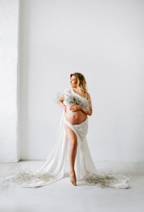A pregnant woman is standing barefoot in a minimalist room with a white background. She is draped in a flowing white cloth and holding a bouquet of delicate flowers. The cloth partially covers her body while some of it spreads on the floor. She has blonde hair that is styled in waves and is looking off into the distance.