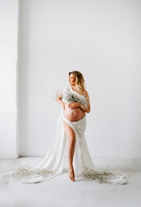 A pregnant woman is standing barefoot in a minimalist room with a white background. She is draped in a flowing white cloth and holding a bouquet of delicate flowers. The cloth partially covers her body while some of it spreads on the floor. She has blonde hair that is styled in waves and is looking off into the distance.