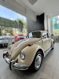 A classic beige vintage car is parked inside a well-lit showroom, with large windows offering a view of a blurred outdoor setting.