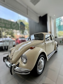 A classic beige vintage car is parked inside a well-lit showroom, with large windows offering a view of a blurred outdoor setting.