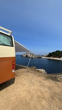 A vintage-style camper van is parked on a dirt patch overlooking a scenic coastal view. The van has an extended awning providing shade. In the distance, a rocky shoreline and lush greenery border the calm, blue ocean under a clear sky.
