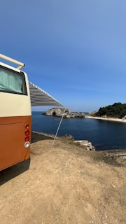 A vintage-style camper van is parked on a dirt patch overlooking a scenic coastal view. The van has an extended awning providing shade. In the distance, a rocky shoreline and lush greenery border the calm, blue ocean under a clear sky.