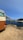 A vintage-style camper van is parked on a dirt patch overlooking a scenic coastal view. The van has an extended awning providing shade. In the distance, a rocky shoreline and lush greenery border the calm, blue ocean under a clear sky.