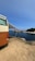 A vintage-style camper van is parked on a dirt patch overlooking a scenic coastal view. The van has an extended awning providing shade. In the distance, a rocky shoreline and lush greenery border the calm, blue ocean under a clear sky.