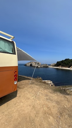 A vintage-style camper van is parked on a dirt patch overlooking a scenic coastal view. The van has an extended awning providing shade. In the distance, a rocky shoreline and lush greenery border the calm, blue ocean under a clear sky.