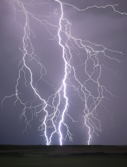 Multiple bright lightning bolts illuminate a dark, cloudy sky, striking down towards an open, flat landscape. The scene is dramatic and vivid, capturing the raw power of a thunderstorm.