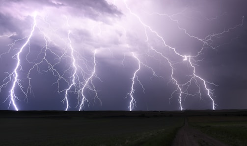 Multiple bolts of lightning illuminate a dark sky over a flat landscape, casting bright, jagged lines across the scene. The ground is dimly visible with a field stretching into the distance.