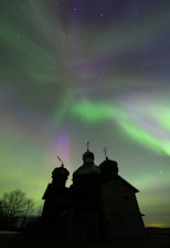 A silhouetted church with domes stands against a vibrant aurora borealis, with green, purple, and hints of pink waves of light stretching across a starry night sky. Bare trees are visible to the left, adding to the serene and captivating atmosphere.