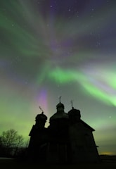 A silhouetted church with domes stands against a vibrant aurora borealis, with green, purple, and hints of pink waves of light stretching across a starry night sky. Bare trees are visible to the left, adding to the serene and captivating atmosphere.