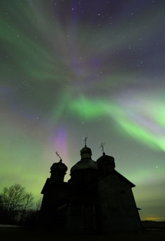 A silhouetted church with domes stands against a vibrant aurora borealis, with green, purple, and hints of pink waves of light stretching across a starry night sky. Bare trees are visible to the left, adding to the serene and captivating atmosphere.