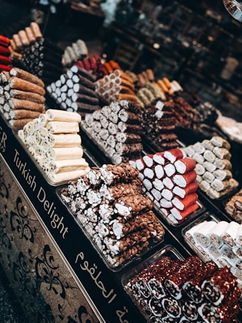 An assortment of Turkish delight is artistically arranged in piles on a market stand. Various types are displayed, including those coated with nuts, powdered sugar, and berry flavors. Each type forms its own colorful stack, creating an inviting and vibrant scene. The stand is adorned with ornate designs and labeled in both English and another language.