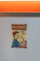 A vintage-style poster depicting a woman with a confident expression, dressed in a blue work shirt and red bandana, reminiscent of the 'Rosie the Riveter' iconography. The woman is flexing her arm to show her strength, with tattoos visible. The poster includes the text 'Warning' and 'Woman at Work', suggesting empowerment and gender equality. The background consists of a tiled wall and an orange panel at the top.